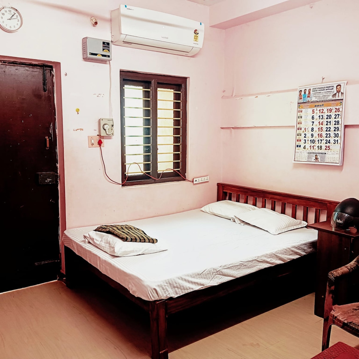 A simple sleeping area is shown featuring a wooden bed with a thin mattress. Sunlight filters through a window with blinds. A calendar is visible on the wall, and a chair is placed in the corner. An air conditioning unit is mounted above the bed.