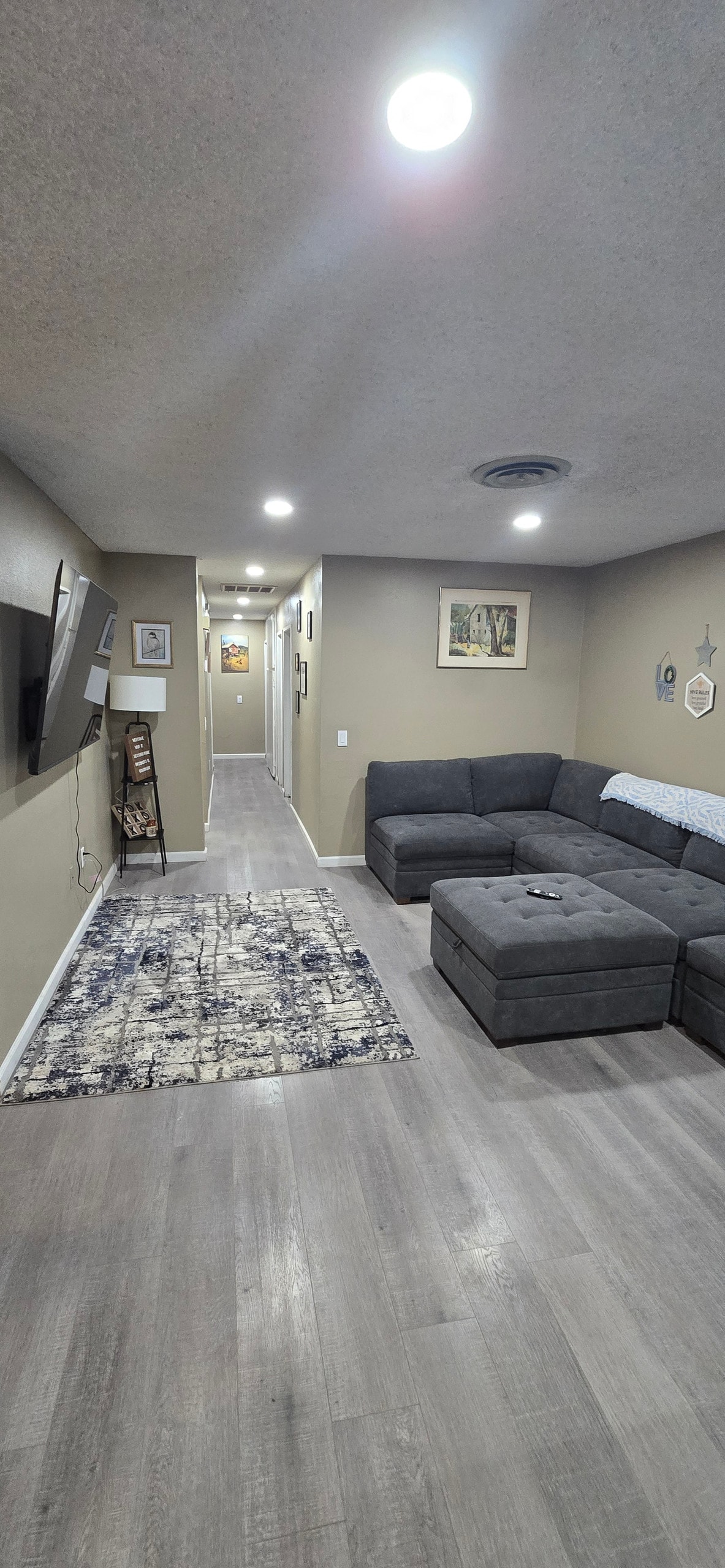 A cozy living area is shown with a large gray sectional sofa positioned against the wall. A television is mounted nearby, and a patterned rug lies on the floor. Soft lighting enhances the inviting space, which leads to a hallway visible in the background.