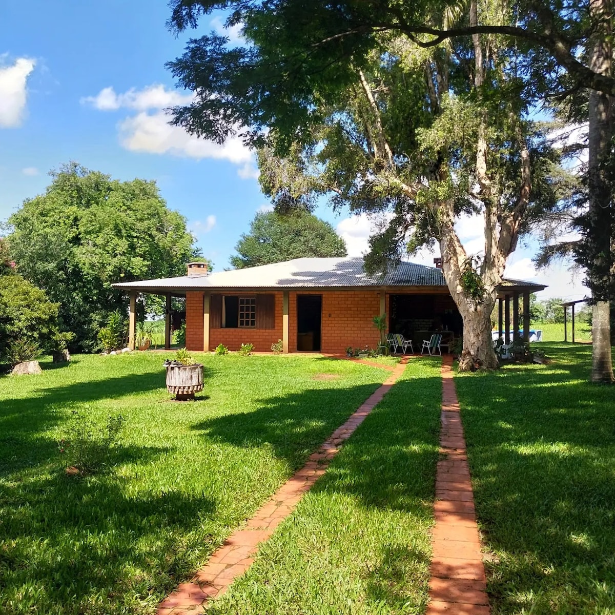 A spacious country house is surrounded by greenery, with a pathway made of brick leading to its entrance. Large trees provide shade, and outdoor seating areas are visible on the porch, highlighting a tranquil and inviting outdoor space.