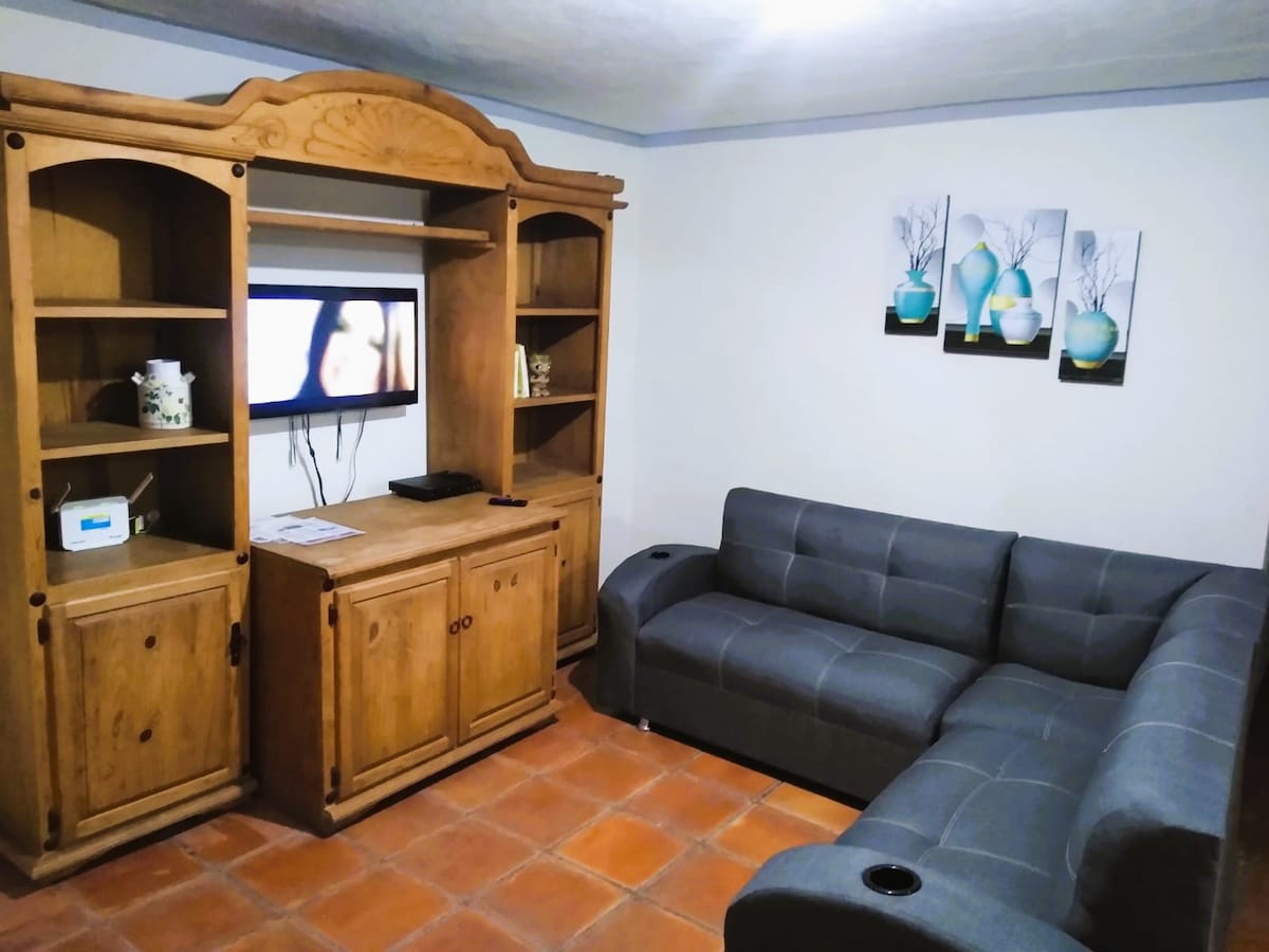 A living area features a wooden shelving unit with a television mounted above it. A gray sectional sofa offers ample seating, while three decorative wall artworks are displayed above it. The floor is tiled, and light enters through a nearby window.
