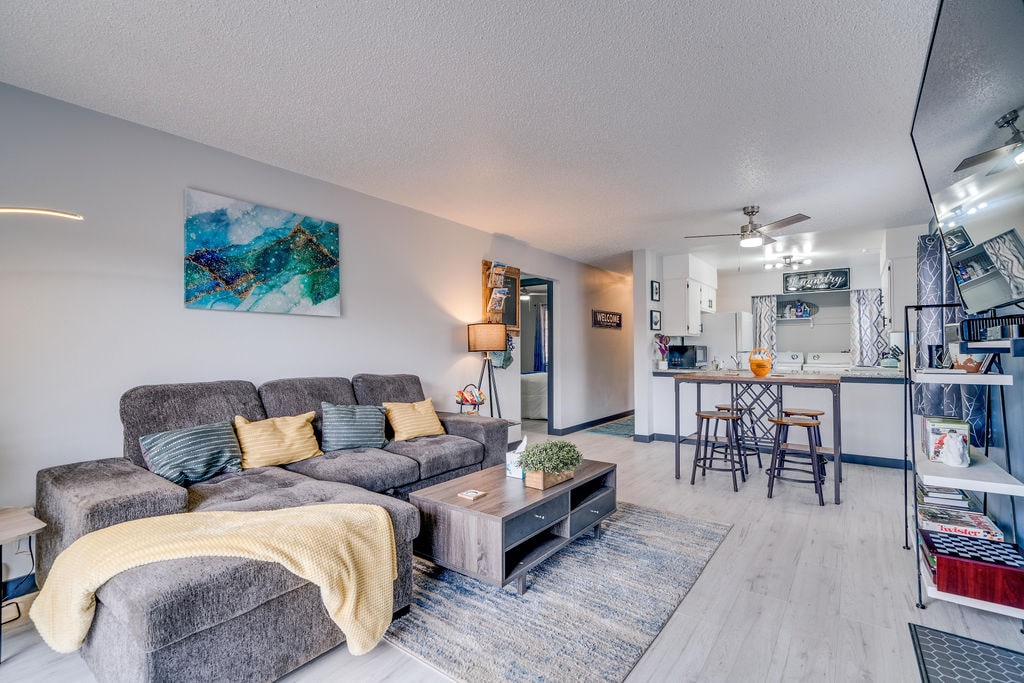 An open living area features a cozy three-seat sofa with a built-in chaise, accompanied by a coffee table. A multi-functional dining space is visible behind, connecting the living area to the kitchen. Natural light brightens the neutral decor and wooden accents throughout the space.