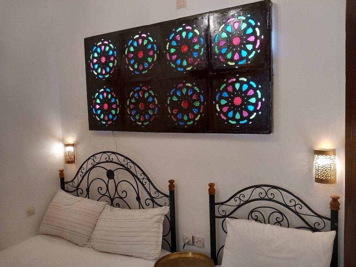 A cozy room features two ornate metal beds with plush white bedding. Above the beds, a decorative wall panel showcases colorful stained glass designs, while softly glowing wall sconces provide gentle illumination. The overall ambiance is enhanced by a clean and simple layout.