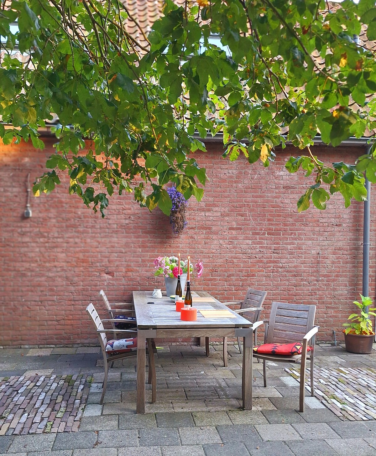 A wooden table is set outdoors under leafy tree branches, surrounded by several wooden chairs. The table is adorned with a bouquet of flowers and bottles, complementing the rustic red brick wall in the background. The cobblestone floor adds to the inviting atmosphere.