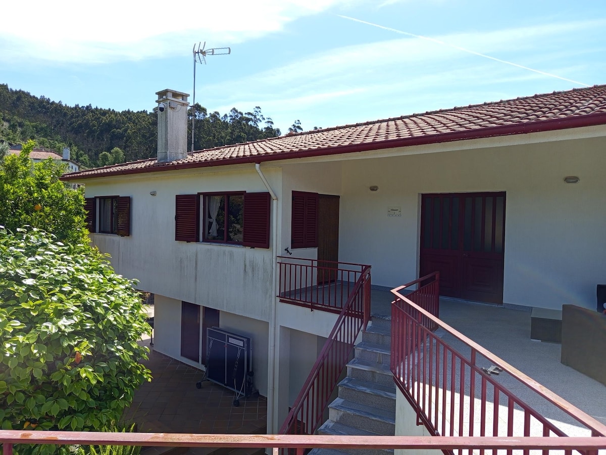 The exterior of the villa features a two-story structure with a sloped roof and red shutters. A staircase leads to an entrance, while outdoor seating is situated on the terrace area. Lush greenery surrounds the property, contributing to a serene atmosphere.