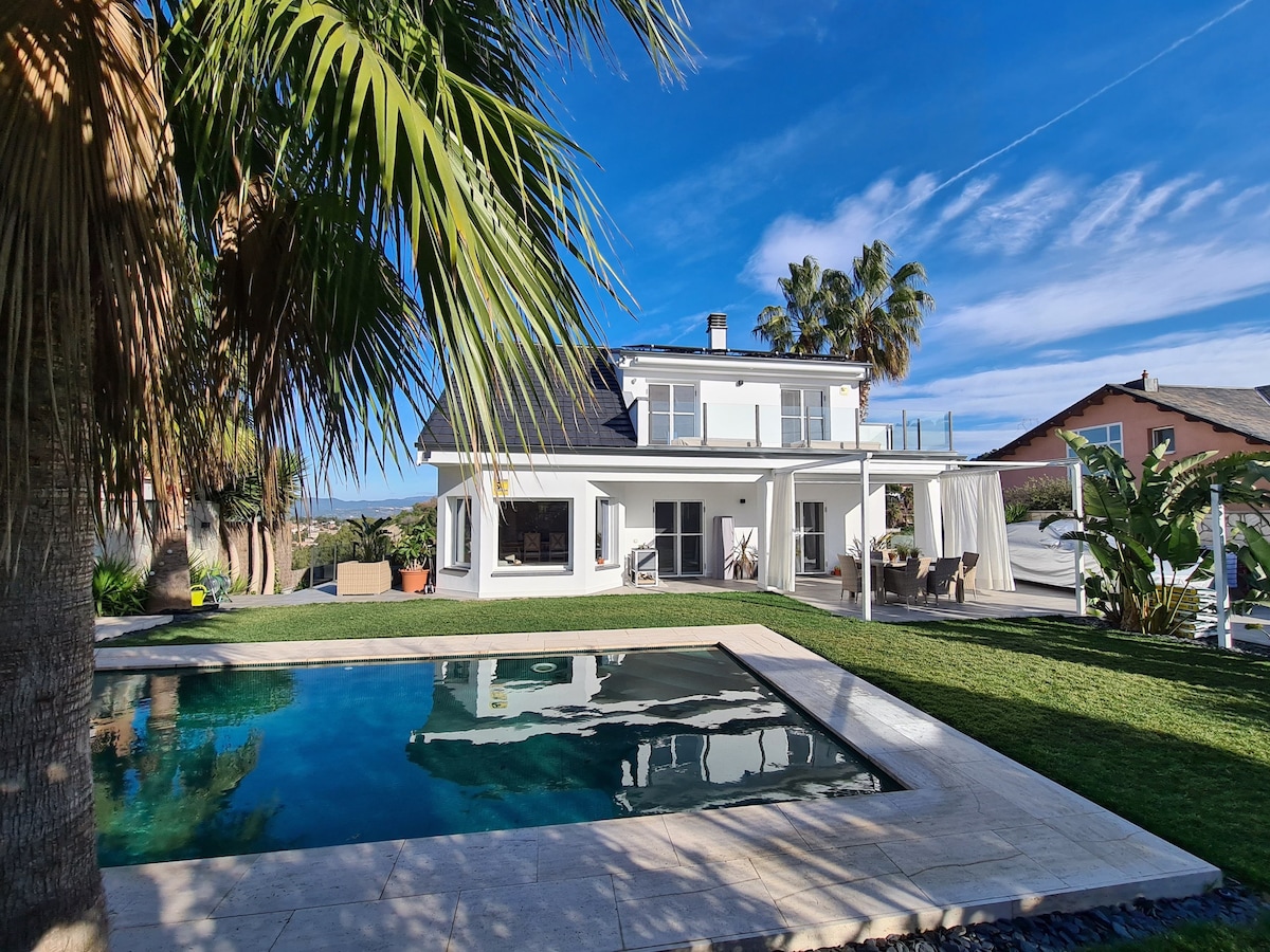 A modern two-story house is surrounded by lush greenery and palm trees. The private pool reflects the clear blue sky, while a spacious patio offers seating. Sunlight illuminates the outdoor area, creating an inviting atmosphere for relaxation and leisure.