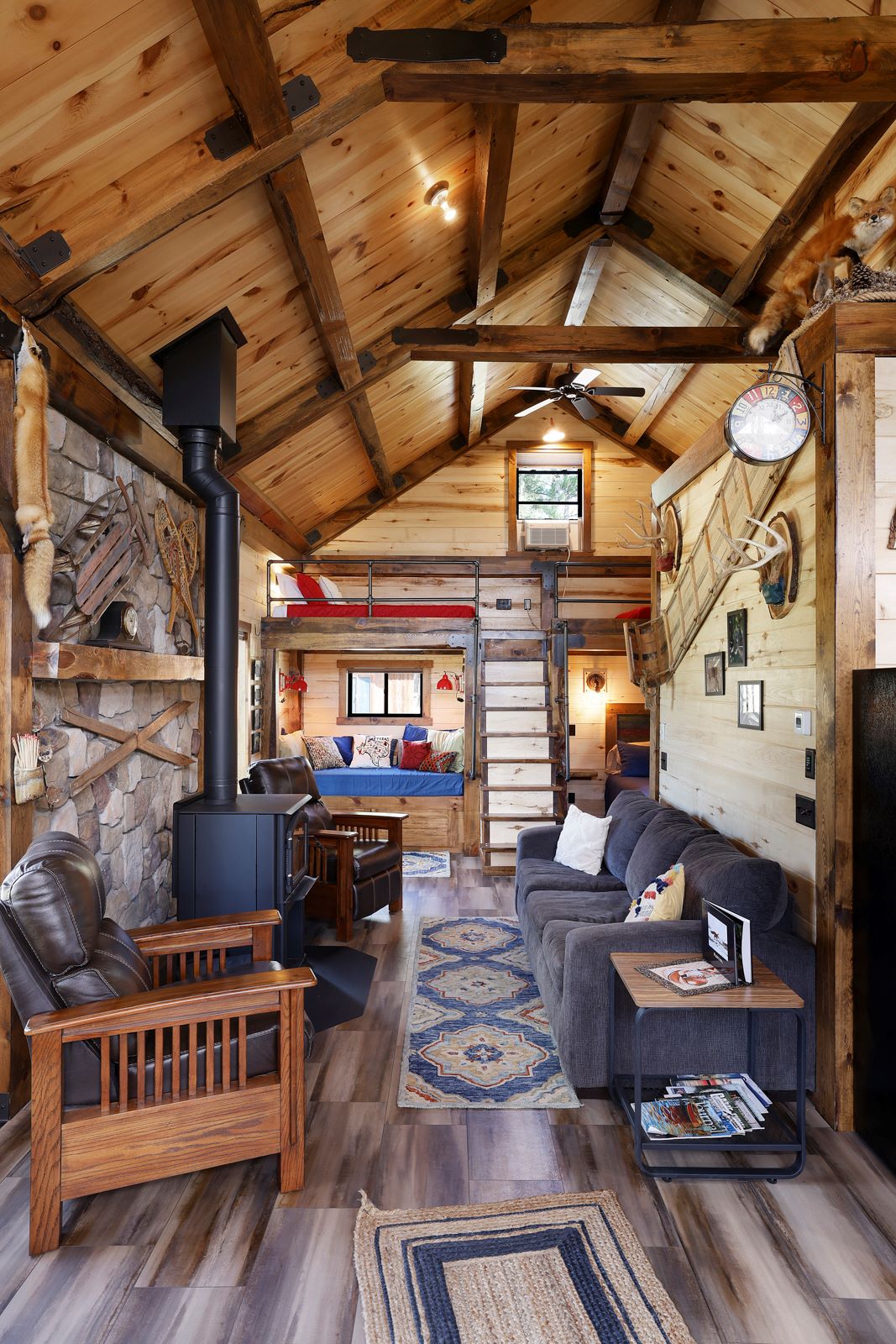 A spacious living area features high, wooden beamed ceilings and a blend of rustic and modern decor. A wood-burning stove sits prominently next to cozy seating, with a patterned rug underfoot. A view of a sleeping area is visible, showcasing a staircase leading to a loft.