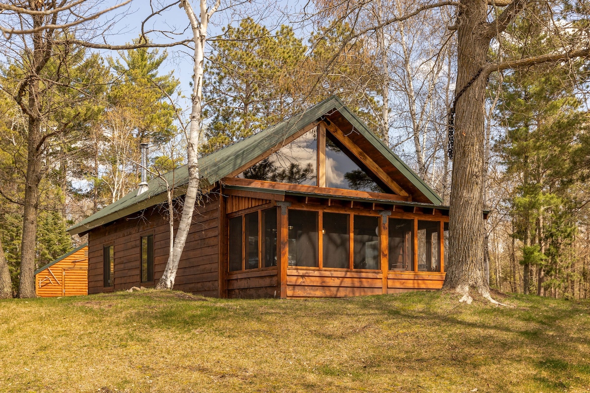 A timber frame cabin is highlighted, featuring a peaked roof and large windows that reflect the surrounding trees. The structure is set back from the lawn, bordered by leafy greenery, providing a sense of seclusion and connection to nature.