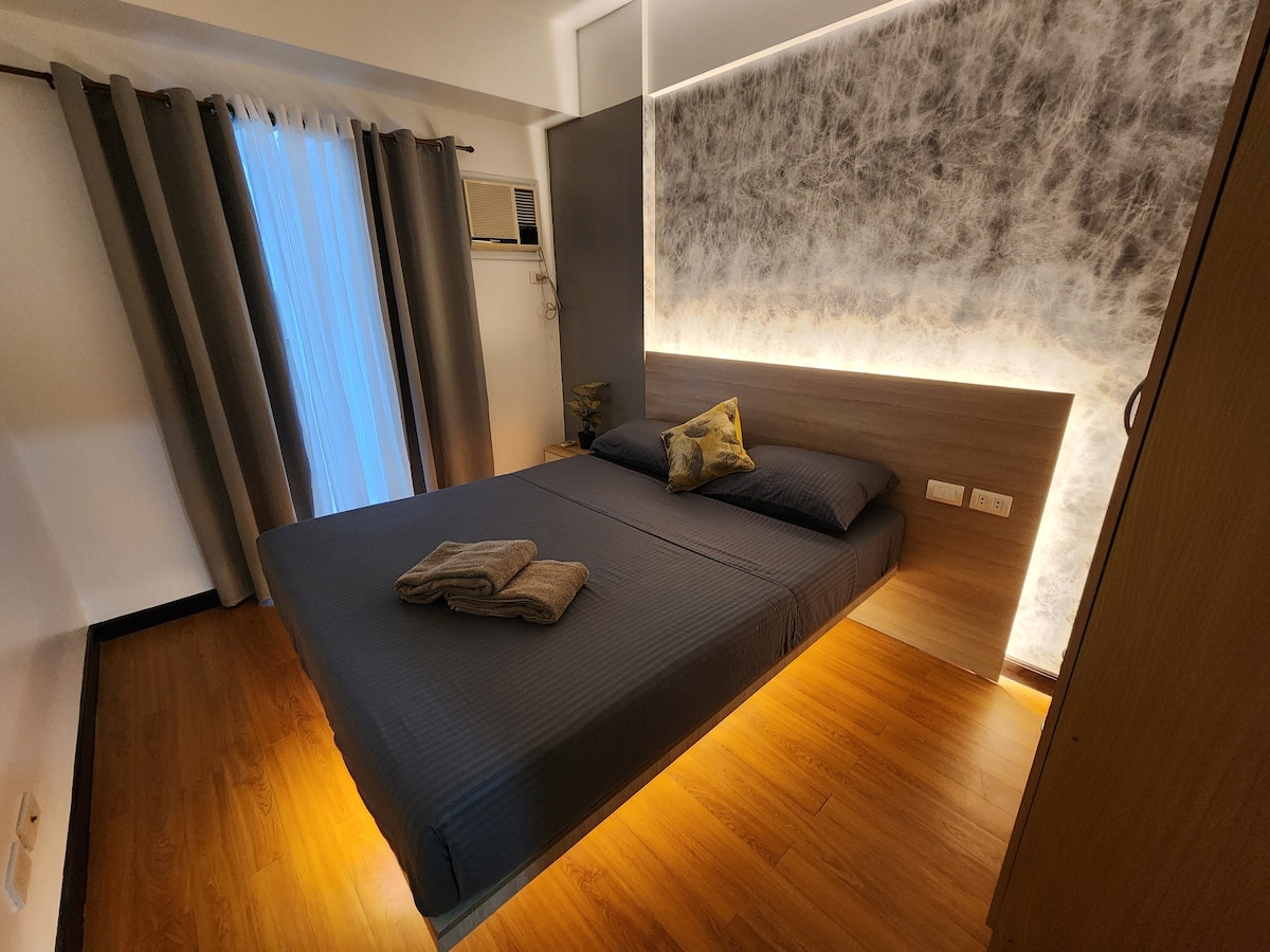 A spacious sleeping area is highlighted by soft lighting along the bed's base. The dark linens complement the light wooden flooring, while floor-to-ceiling curtains frame a window to enhance natural light. A decorative pillow sits on the neatly made bed.