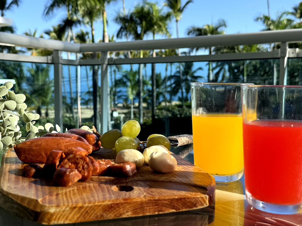 A wooden platter displays an assortment of local fruits and snacks, accompanied by two glasses of brightly colored beverages. The background features a balcony with tropical palm trees and a glimpse of the ocean, enhancing the inviting ambiance.