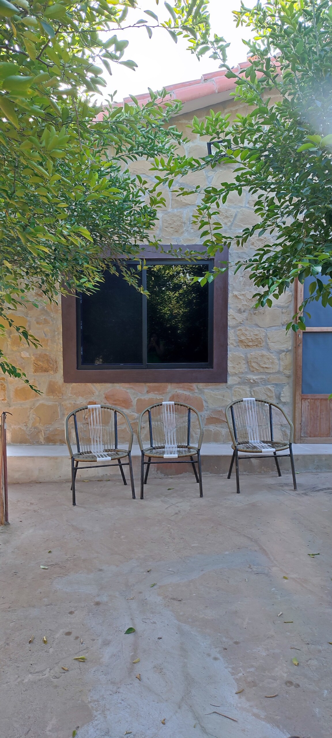 Three chairs are arranged outside a stone wall, positioned in front of a large window. Lush greenery surrounds the area, offering a natural setting. The ground is composed of earthy tones, complementing the warm stone features of the building.