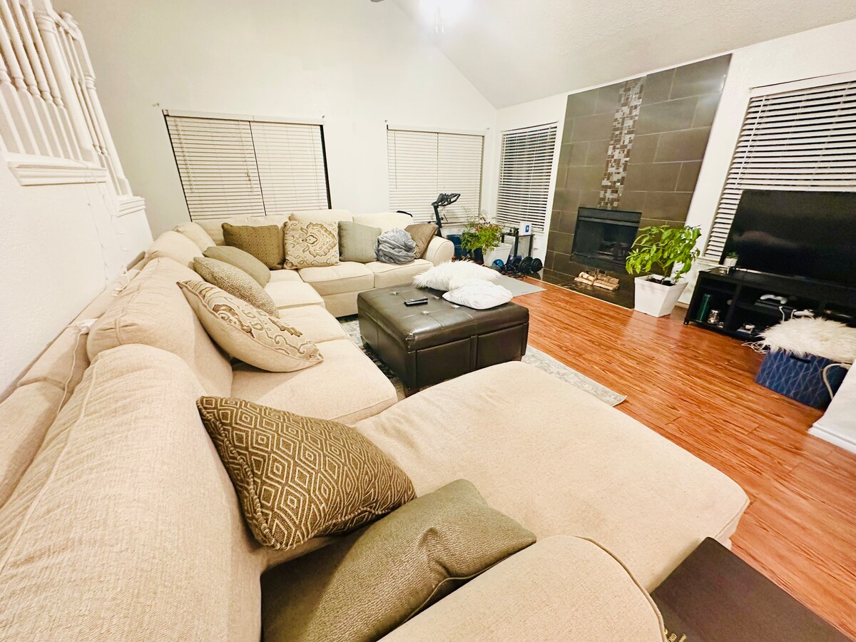 A spacious living room features a large sectional sofa adorned with decorative pillows. A flat-screen television is positioned next to a modern fireplace, and potted plants add greenery to the area. Natural light is allowed in through multiple windows with blinds.