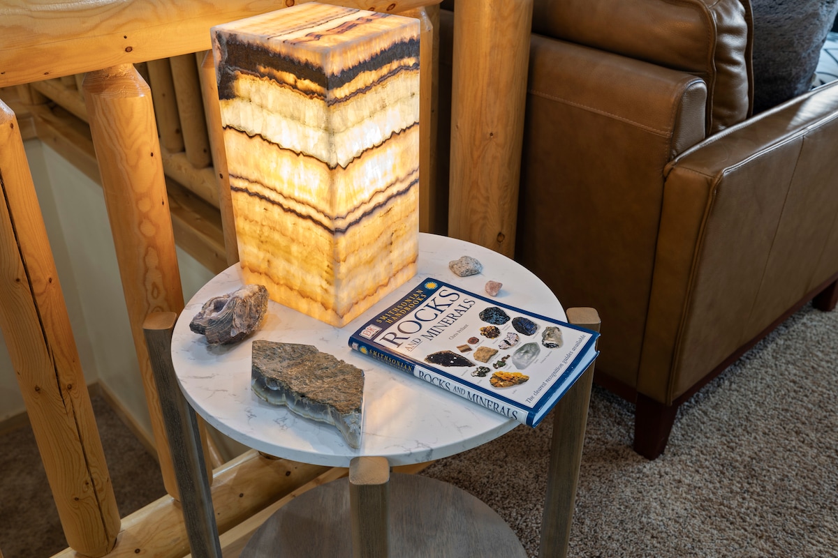 A side table features a decorative onyx lamp, radiating warm light. A geology book rests on the table, surrounded by various natural stones and minerals, offering a touch of rustic charm. The setting is complemented by wooden structure details in the background.