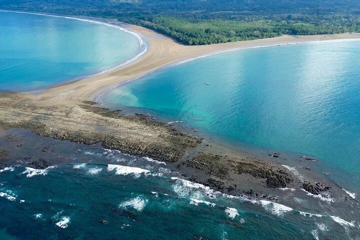 Aerial view of a serene coastline reveals a curve of sandy beach meeting tranquil turquoise waters. Rocky formations emerge from the ocean, creating natural contrasts. Lush greenery lines the shore, enhancing the scenic landscape while waves gently lap against the sand.