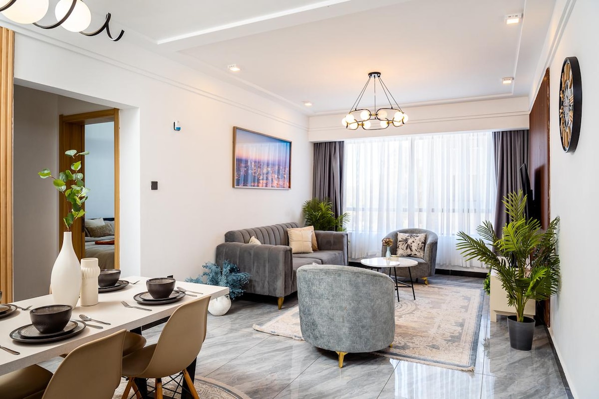 The living area features a cozy gray sofa and armchairs, complemented by a round coffee table. A dining table set is visible, adorned with tableware. Natural light fills the space through large windows, adding a refreshing touch to the modern decor.