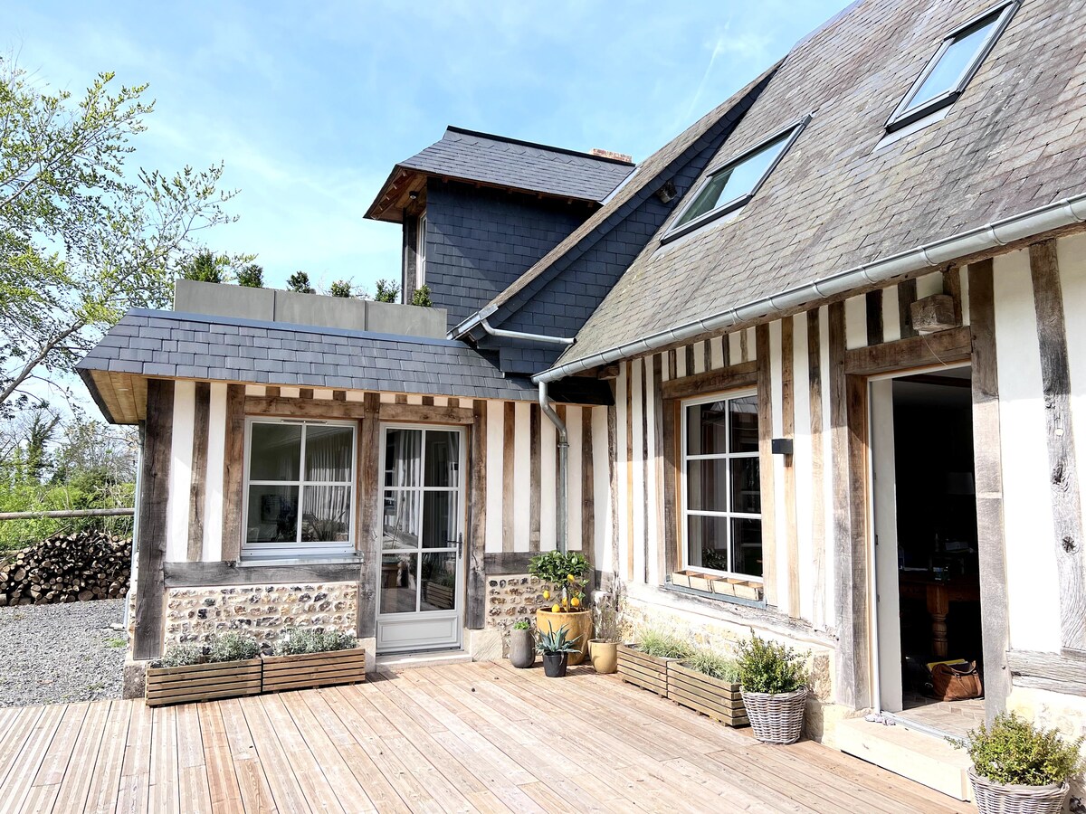 The exterior of a charming house features a wooden deck leading to a set of large windows framed by traditional wood paneling. The roof is adorned with greenery, and potted plants are placed along the deck, creating a connection to the surrounding nature.