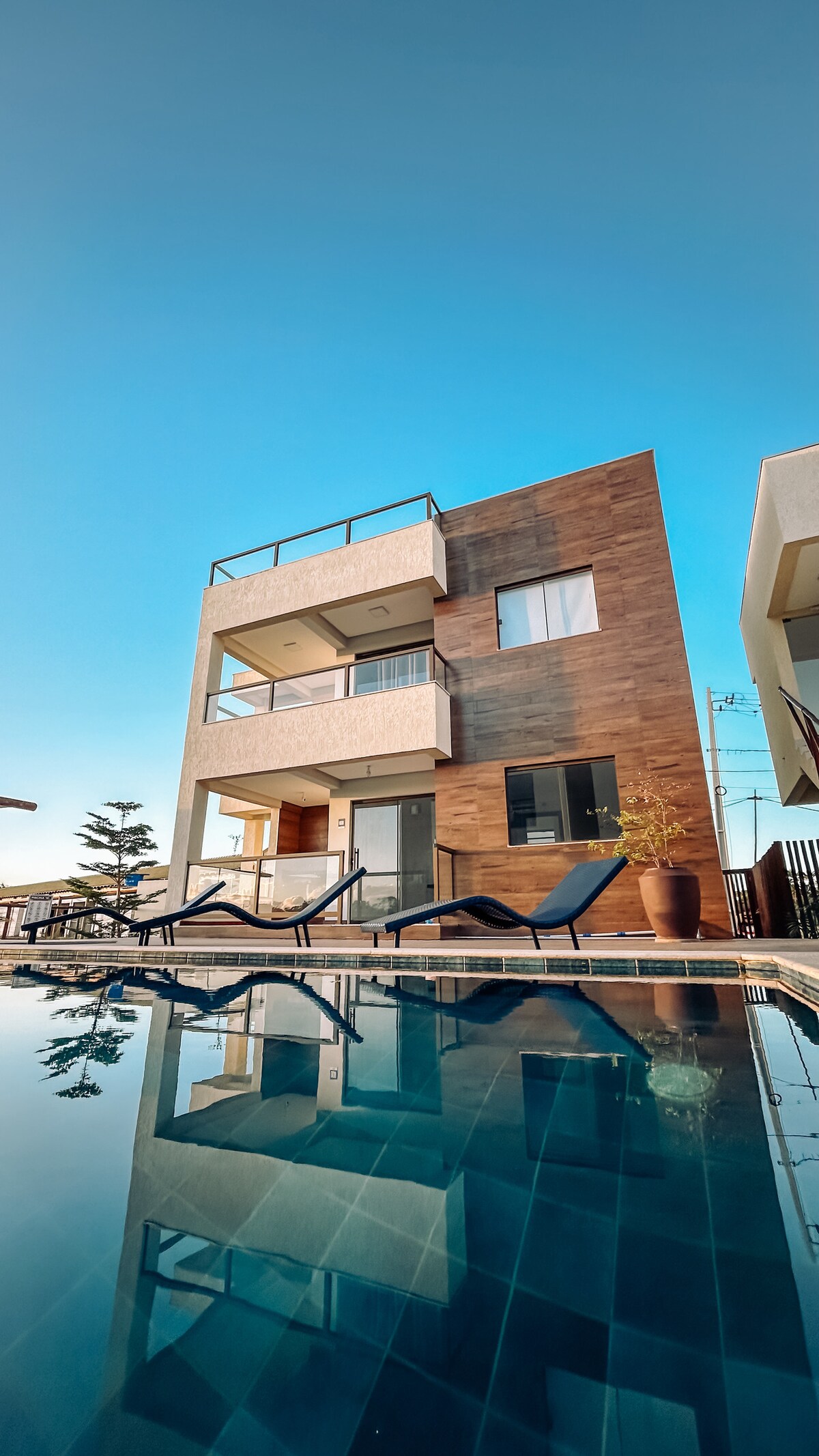A modern three-story building stands against a clear blue sky, reflecting in the tranquil pool below. Lounge chairs surround the pool, and a lush potted plant adds a touch of greenery, enhancing the inviting outdoor space.