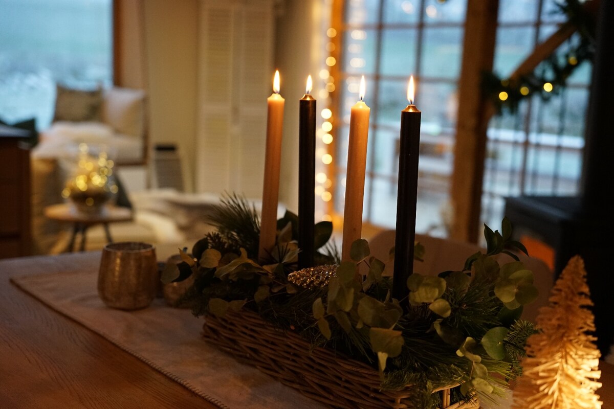 A cozy interior setting features a decorative centerpiece with four candles and greenery, placed on a wooden table. Soft lighting from nearby sources casts a warm glow, enhancing the inviting atmosphere. The background includes a comfortable seating area and large windows with scenic views.