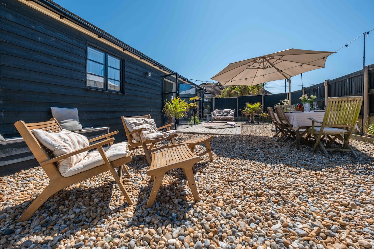 An outdoor area is presented with pebbled flooring and wooden furniture, including four lounge chairs positioned around a small table. A large umbrella provides shade over a dining table surrounded by wooden chairs, creating a relaxed space for dining and gatherings.