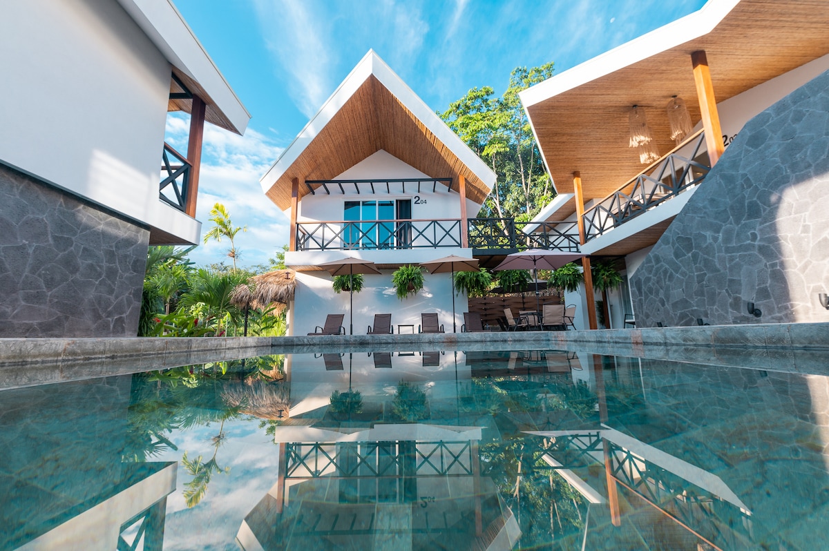 The image showcases a serene outdoor setting with a jade-colored pool reflecting the architectural elements of the two-story retreat. Tropical greenery and wooden accents frame the property, inviting relaxation and connection with nature under a bright blue sky.