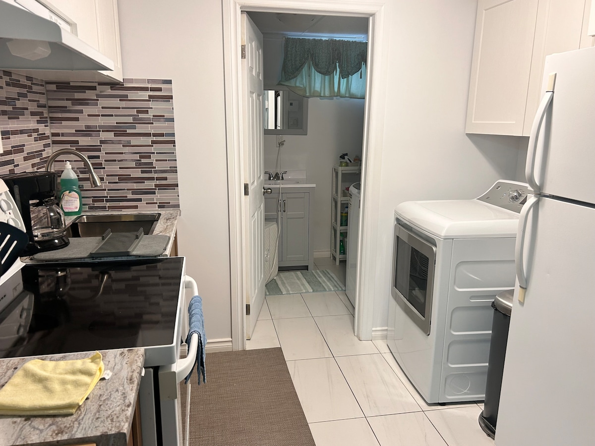 A fully equipped kitchen is seen featuring modern appliances, including a stove and refrigerator. The layout offers a view into a nearby bathroom with laundry facilities. Light-colored cabinets and a decorative backsplash provide an appealing look, complemented by ceramic tile flooring.