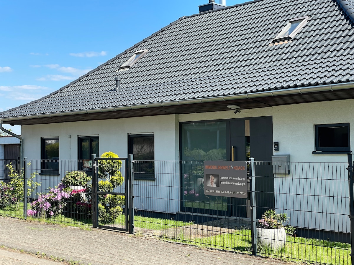 A modern two-family house is viewed from the front, featuring a neatly maintained garden with shrubs and flowering plants. A gated entry leads to the main entrance, which is highlighted by large glass doors. The roof is adorned with traditional tiles against a clear blue sky.