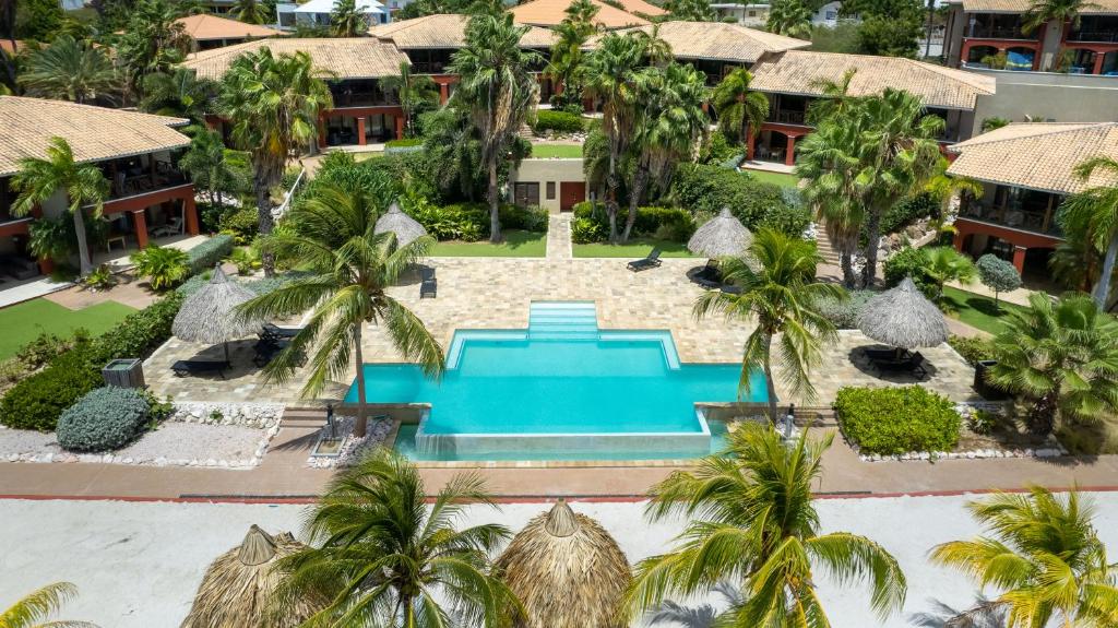 A lushly landscaped resort is presented, featuring a central swimming pool surrounded by palm trees and lounge chairs. Thatched-roof cabanas offer shade, and pathways connect the various buildings in the complex, showcasing a tranquil outdoor environment.