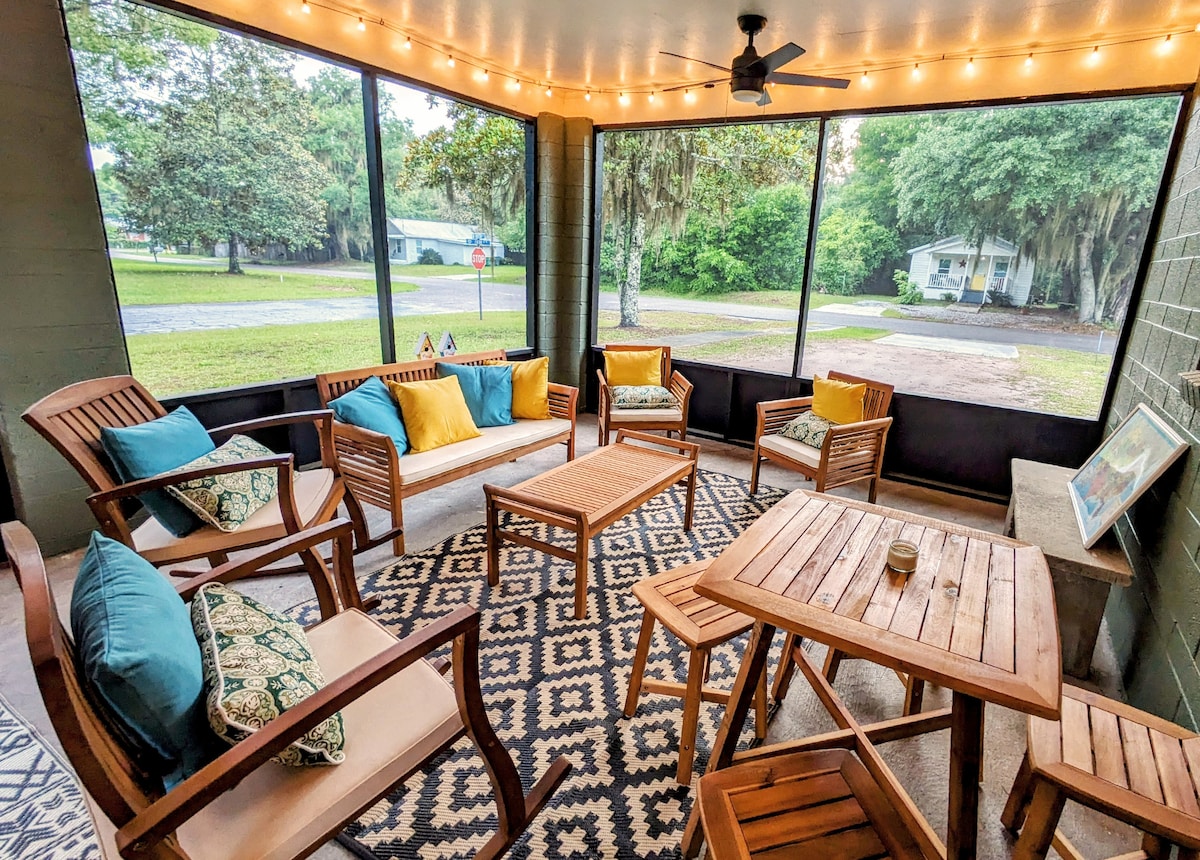 A screened-in patio is furnished with a mix of comfortable seating, including two chairs, a couch, and a table. Decorative pillows add color to the space, which is enhanced by warm string lights overhead. A patterned rug complements the outdoor atmosphere.