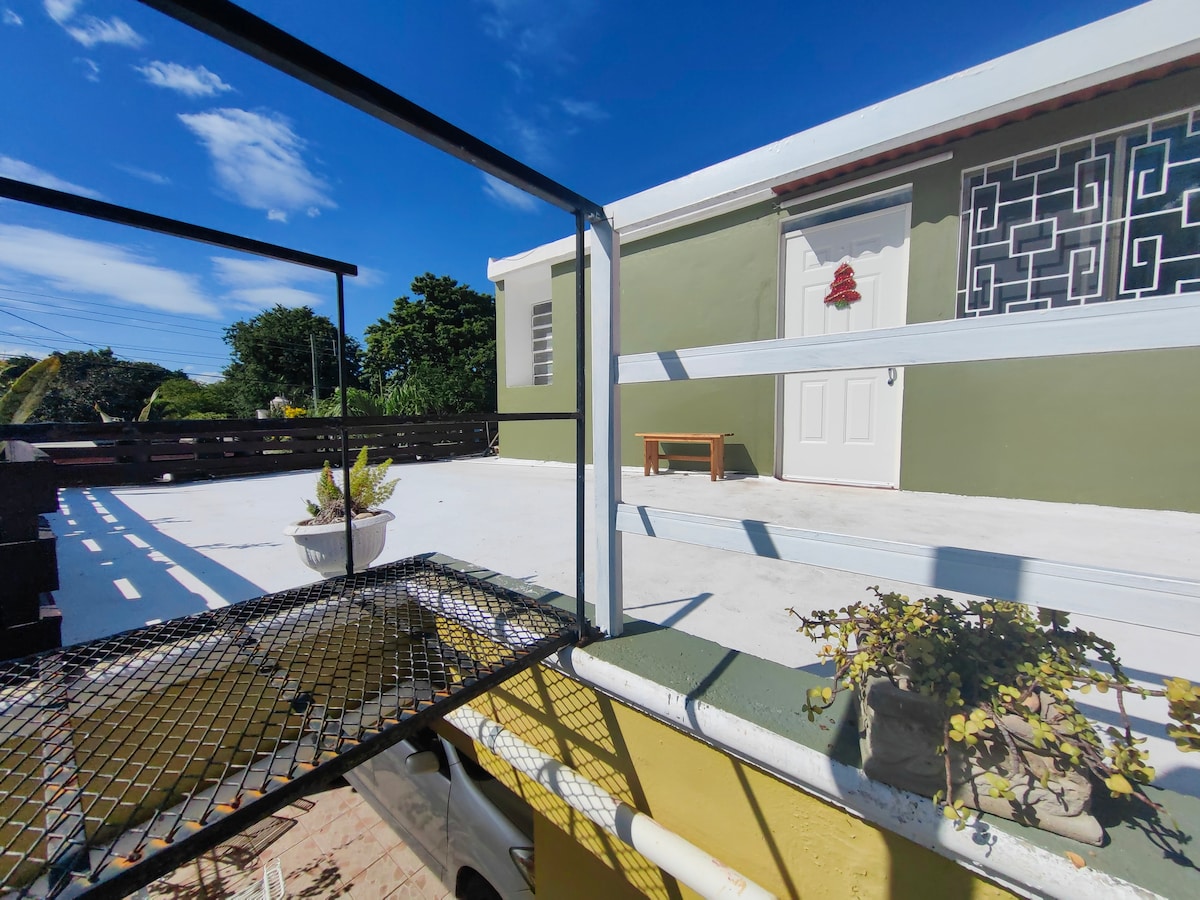 An exterior view features a staircase leading up to the second floor of the property. A landing area is visible with a potted plant and a wooden bench beside a door. The sky is clear, enhancing the appearance of the surrounding greenery.