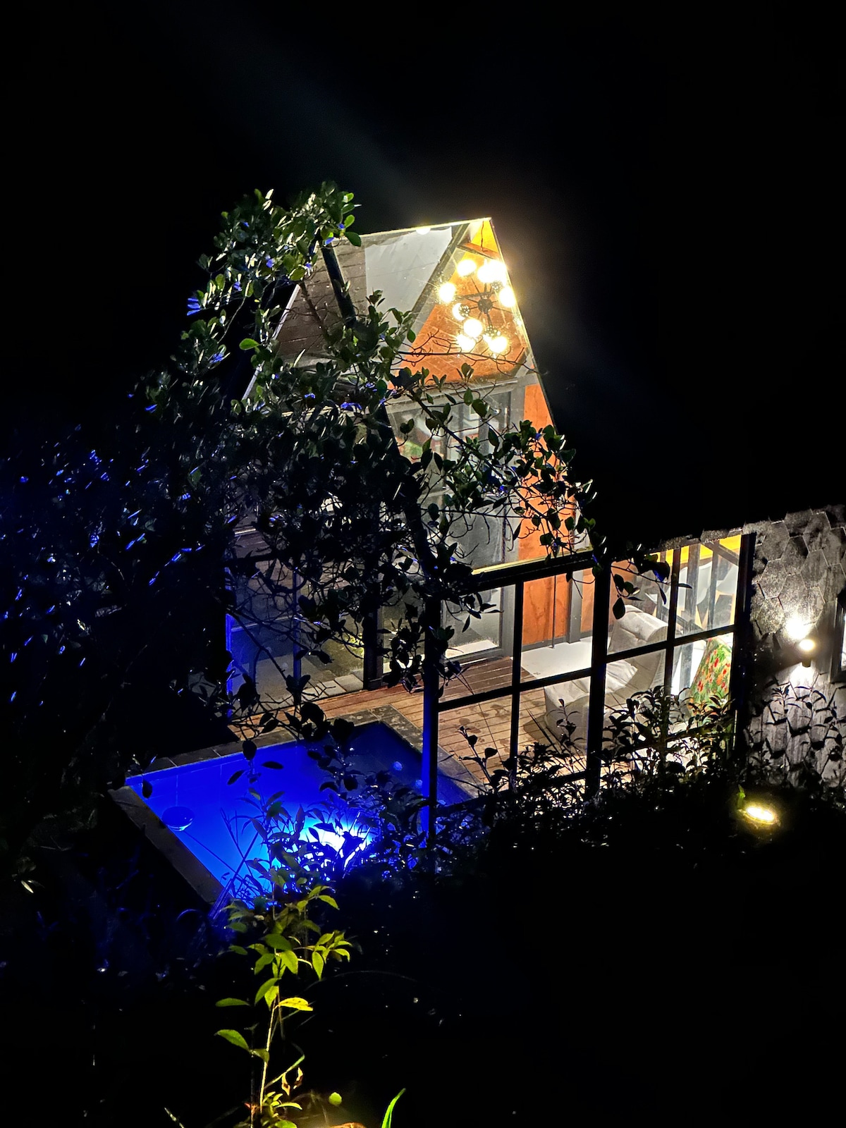 The cabin is illuminated at night, showcasing its unique architectural design with a sloped roof. Soft lighting highlights the outdoor area and adjacent pool, while surrounding vegetation adds a natural touch to the tranquil setting.