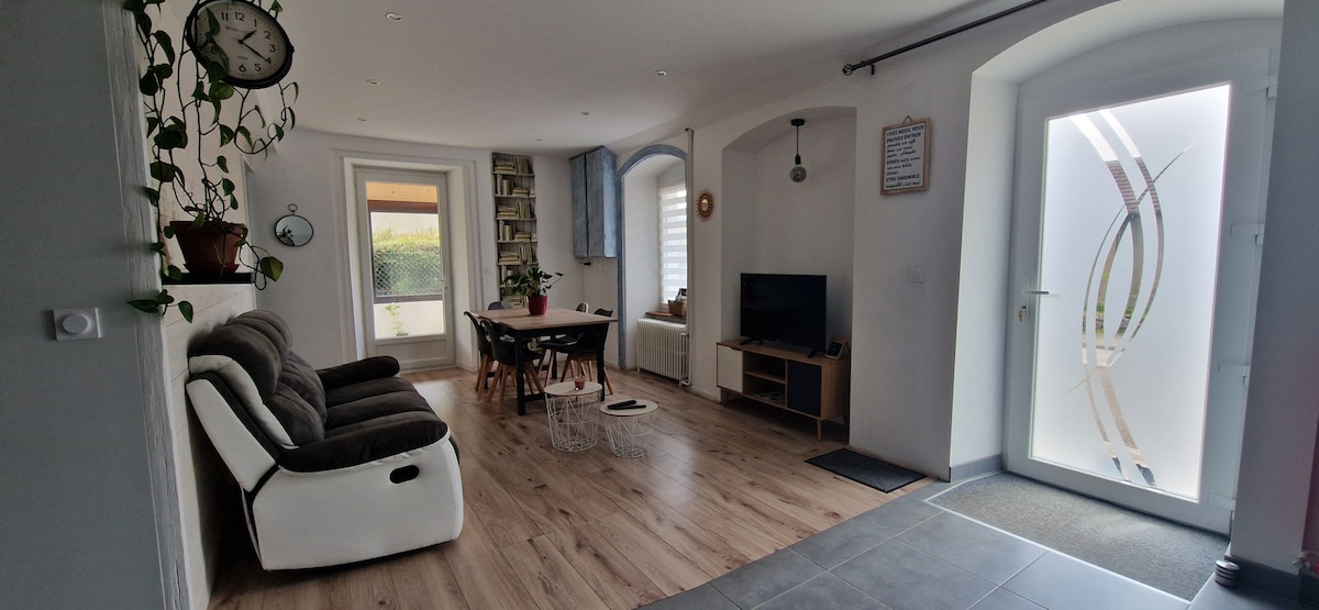 An open living area features a comfortable seating arrangement around a wooden dining table. Natural light floods through large windows, with a view of the surrounding greenery. Decorative elements include a wall clock and potted plants, offering a sense of tranquility.