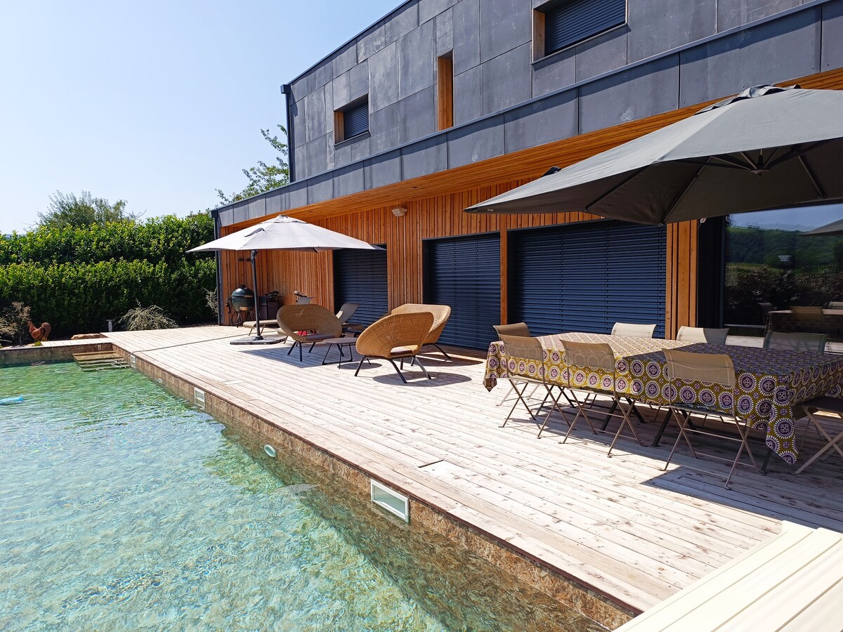 A spacious wooden terrace features a partially shaded dining area complemented by patterned tablecloths. Sun loungers are arranged beside a clear pool, reflecting the blue sky. Umbrellas provide shade, creating a comfortable setting for relaxation and outdoor dining.