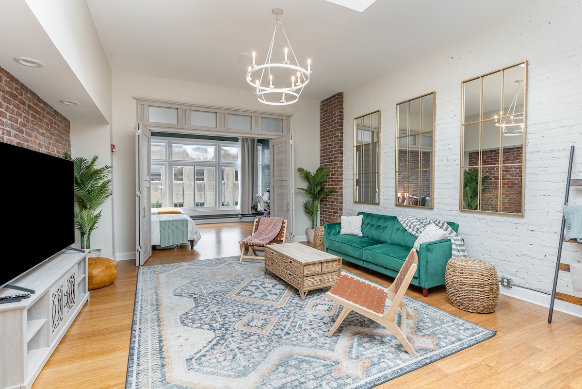 A spacious living area features a green sofa and decorative chairs. A large area rug covers the hardwood floor. Mirrors adorn the walls, reflecting natural light streaming through the expansive windows. Decorative plants are placed in the corners, enhancing the room's open feel.