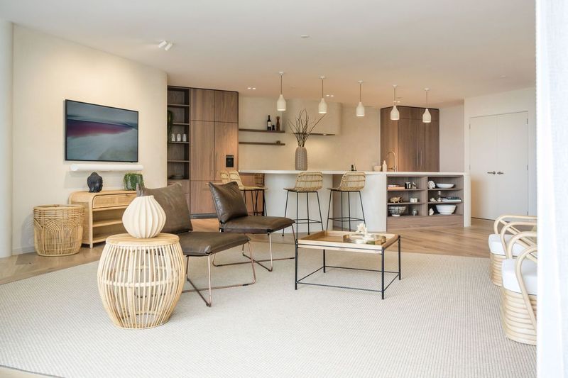 A modern living area features a combination of natural textures and muted tones. A spacious seating arrangement includes a dark leather chair and a woven coffee table. The kitchen area, equipped with stylish bar stools, showcases minimalistic cabinetry and light fixtures.