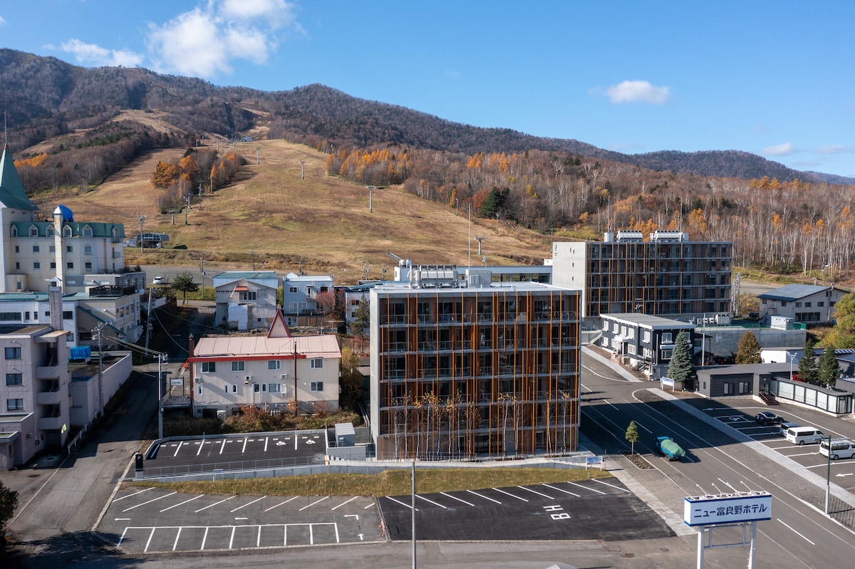 An aerial view of Fenix West is presented, showcasing its modern architecture surrounded by open spaces. In the background, the Tokachi mountain range is visible, with autumn foliage adorning the landscape. A parking area is neatly arranged in front of the condominium.