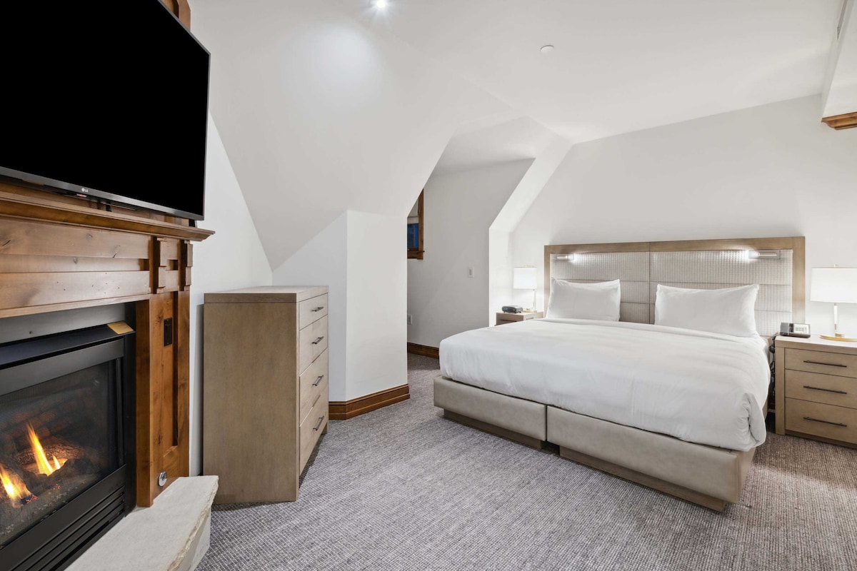 A spacious sleeping area is highlighted by a king size bed with soft bedding and pillows. A cozy fireplace is positioned to one side, while a flatscreen TV is mounted above a wooden dresser, offering a modern touch to the room's welcoming ambiance.