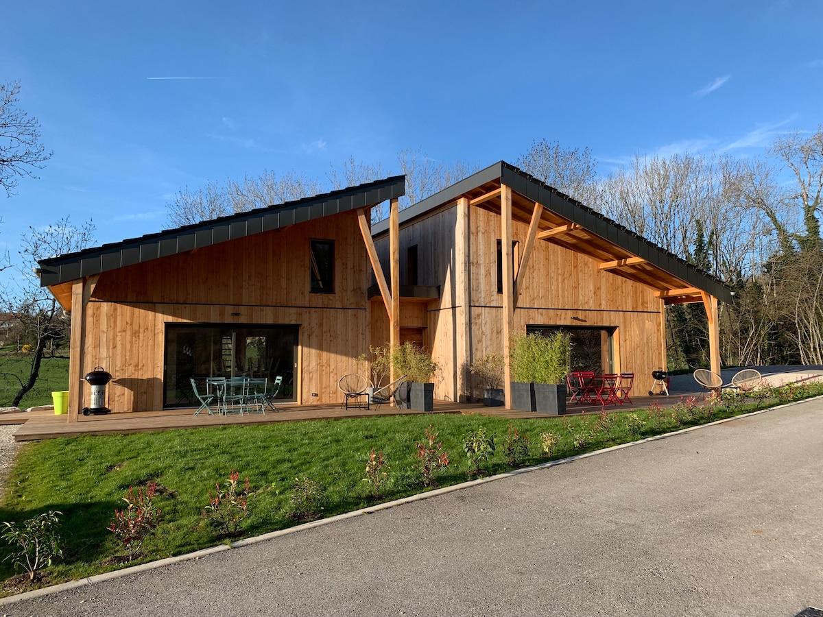 A modern wooden lodge is showcased, featuring a unique architectural design with slanted roofs. Large windows provide ample natural light, while a spacious terrace extends out front. Colorful outdoor furniture, including tables and chairs, complements the landscaped grassy area.