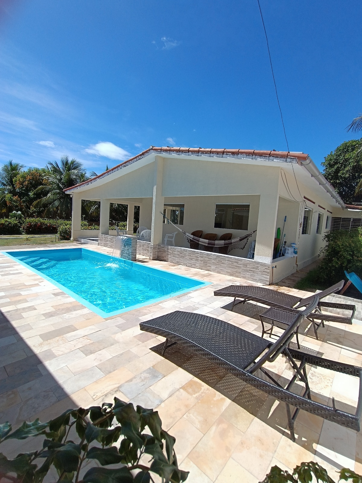 The image showcases a single-story house with large windows, surrounded by a well-maintained garden. A clear pool is visible in the foreground, complemented by two reclining lounge chairs. Lush greenery and palm trees provide a tropical backdrop under a bright blue sky.