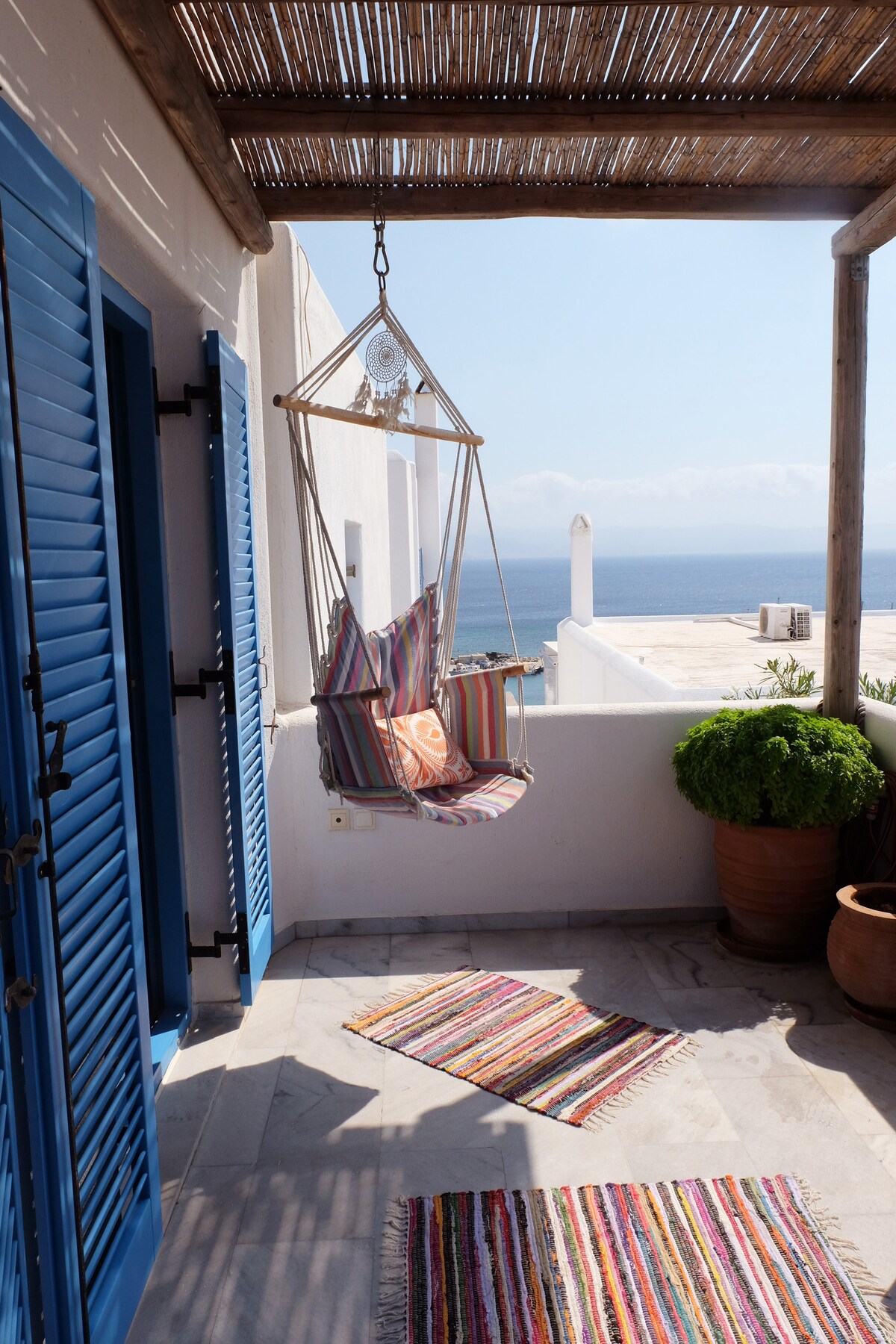 A charming outdoor space is depicted featuring a colorful hanging chair suspended from a wooden frame. A striped rug lays on the marble floor, complemented by potted greenery. The tranquil sea view is visible, framed by light, airy walls and vibrant blue shutters.