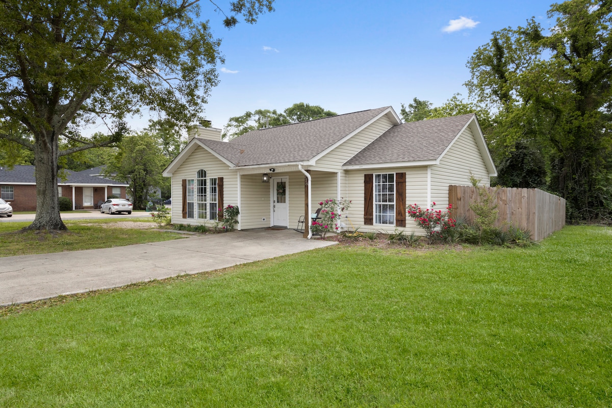 A charming single-story home is set on a grassy lawn, framed by trees. The exterior features light-colored siding with two front windows and a covered entryway. Vibrant flower beds are positioned near the entrance, and a wooden fence provides privacy in the backyard.