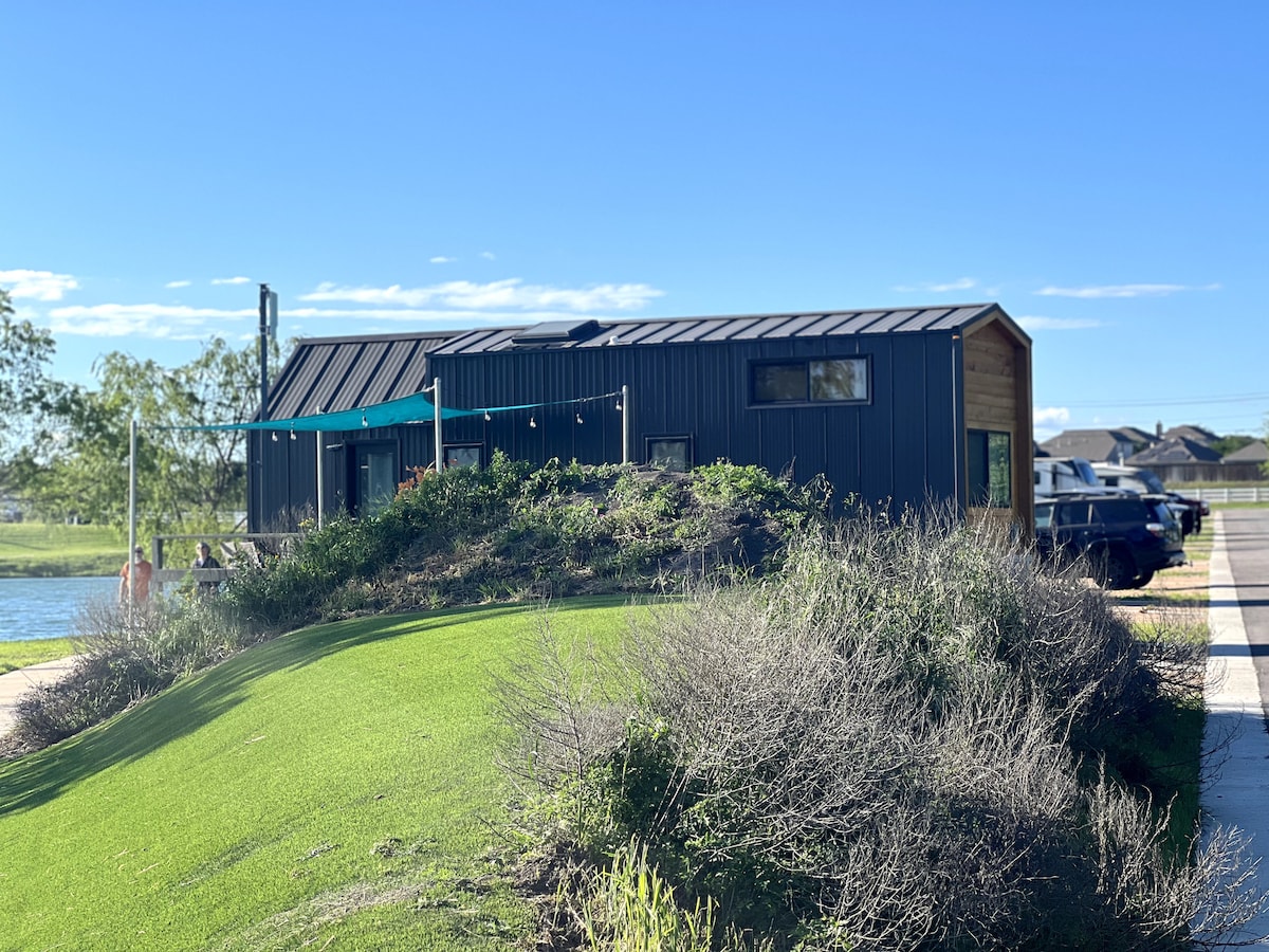 The Tiny House is positioned next to a serene lake, nestled against a gentle hill. A patio extends from the home, adorned with overhead string lights. Green grass and shrubs surround the base, creating a welcoming outdoor space for relaxation.