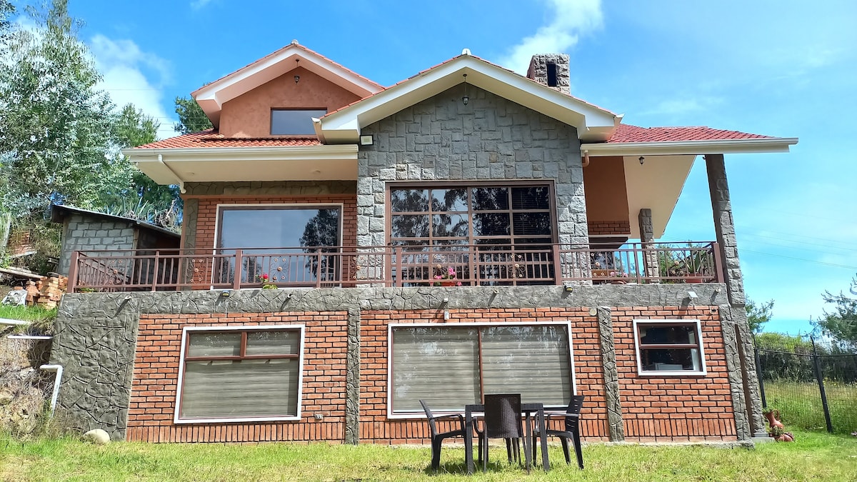 The exterior of a spacious two-story building is shown, featuring a mix of stone and brickwork. Large windows allow for ample natural light. A small patio area with a table and chairs is visible in front, surrounded by greenery and under a clear blue sky.