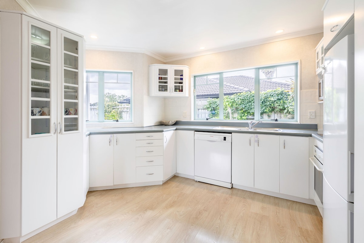 A modern kitchen is designed with white cabinetry and a light countertop. Multiple windows allow natural light to fill the space, while a dishwasher and other appliances are neatly integrated. The flooring is a light wood, enhancing the kitchen's airy feel.
