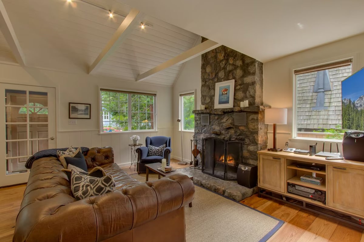 A cozy living room features a stone fireplace with a warm flame. A leather couch is positioned to provide comfort, complemented by a large area rug. Natural light filters through multiple windows, illuminating the space with a welcoming ambiance.