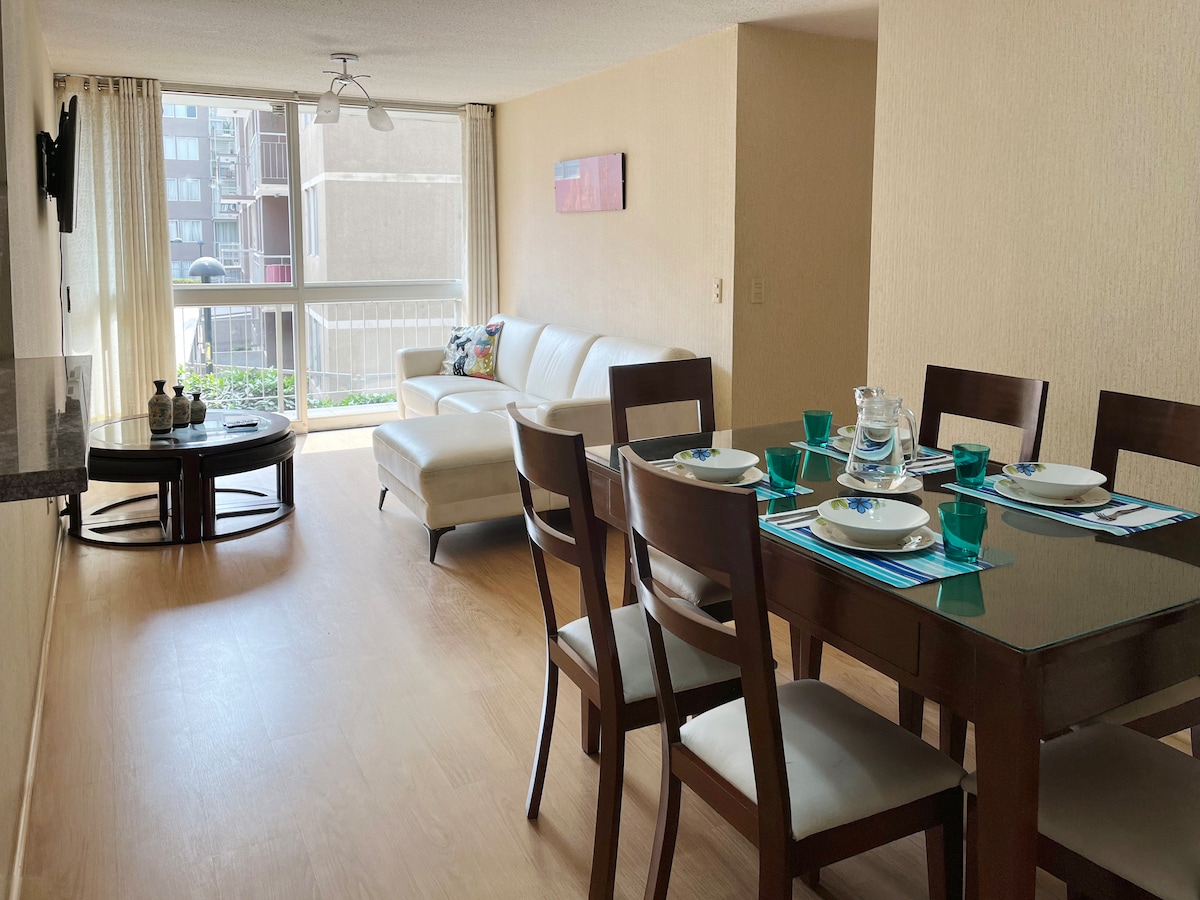 A bright living and dining area features a spacious white sofa and a round coffee table with decorative items. A dining table is set for six, adorned with teal dishware. Floor-to-ceiling windows provide ample natural light, enhancing the openness of the space.