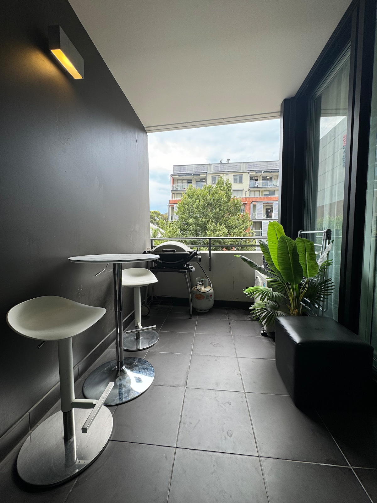 A small balcony is presented with sleek dark tiles and a round table accompanied by two stools. A potted plant adds greenery, while an outdoor grill is positioned nearby, enhancing the functionality of the space.