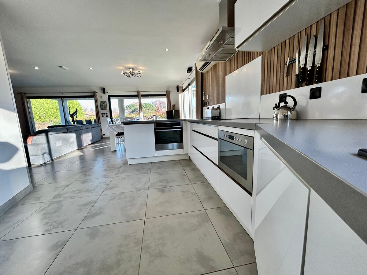 A modern kitchen is presented with sleek cabinetry and stainless steel appliances. A spacious countertop allows for meal preparation, while the open layout leads to a living area with large windows. Natural light brightens the space, highlighting the tiled flooring.