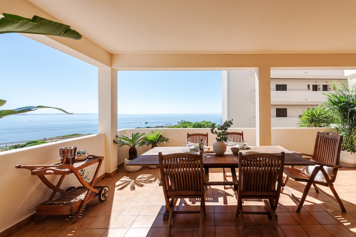 An outdoor terrace offers a spacious wooden dining table surrounded by several wooden chairs. Potted plants and a cart are visible, while panoramic views of the sea and clear blue sky create a serene backdrop.