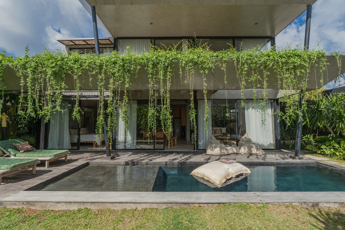 A spacious villa is surrounded by lush greenery, featuring a private swimming pool in front. The pool is bordered by wooden decking and plush lounging cushions are placed nearby. Floor-to-ceiling glass doors offer a view into the airy interior spaces of the villa.