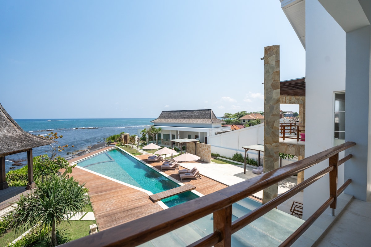 An expansive view captures the serene infinity pool blending seamlessly with the ocean. Sun loungers are arranged along the deck, and shaded seating areas provide relaxed spaces. The villa's modern design is complemented by landscaped gardens, with the ocean horizon extending in the background.