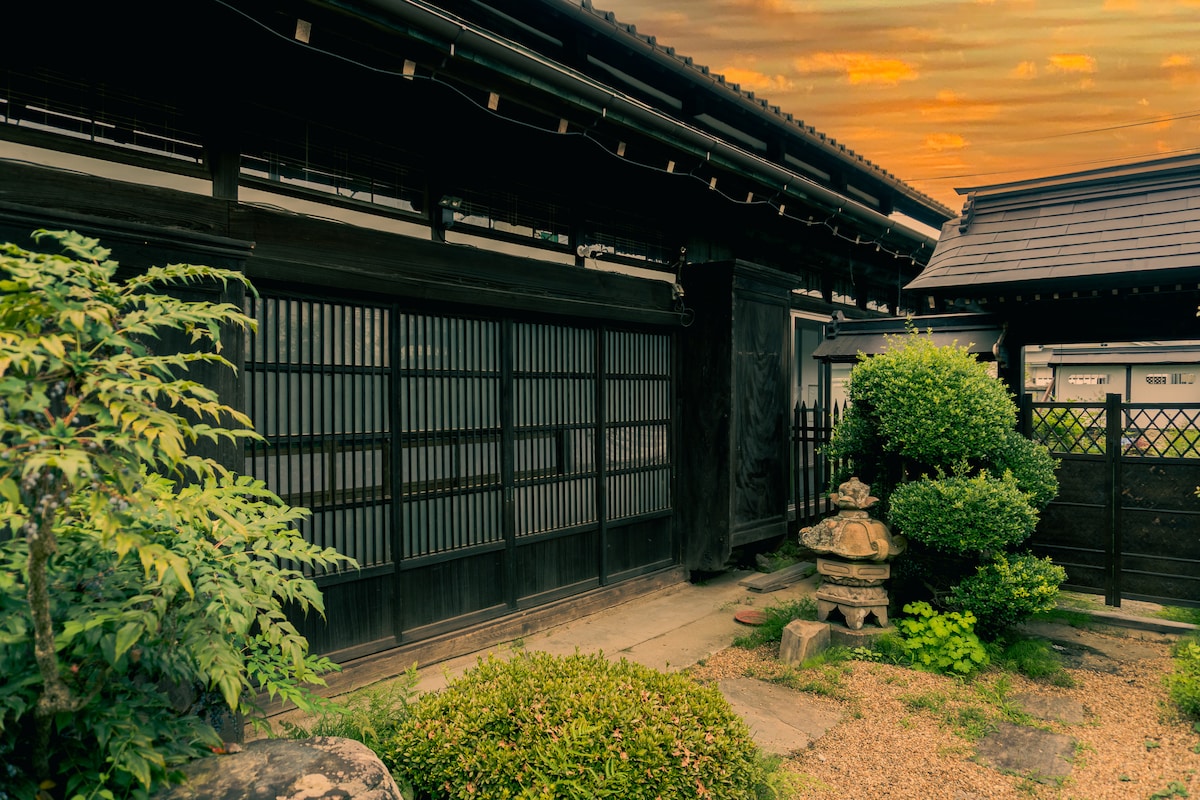 The exterior of a traditional Japanese farmhouse is showcased, featuring wooden siding and shoji screens. Lush greenery surrounds the structure, with neatly trimmed bushes and a stone lantern in the garden. A warm sunset sky adds a serene ambiance to the scene.